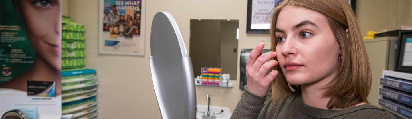 a woman trying on a contact lens and a hand mirror spokane eye clinic